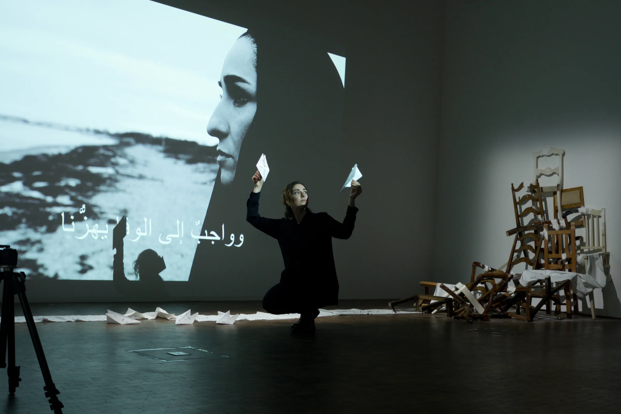 Areej Almansory kneels in a darkened gallery holding two folded paper forms, one raised in each hand, with a large projected photograph and Arabic text behind her and a stack of broken wooden chairs to the right.