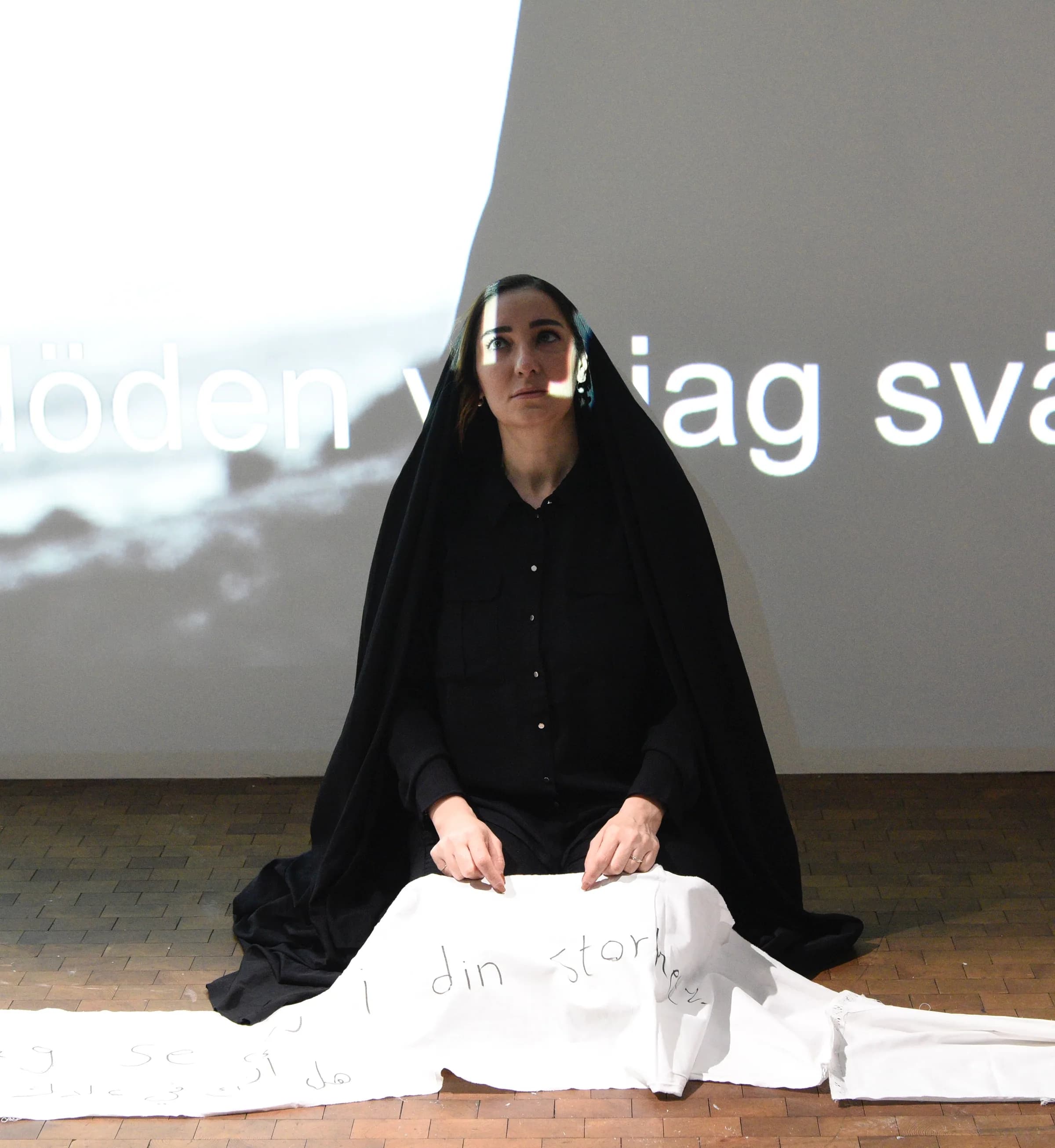 During a performance, a woman draped in a long black cloth sits on the floor holding a white textile in her lap and looks upward; large projected Swedish text runs across the wall behind her.