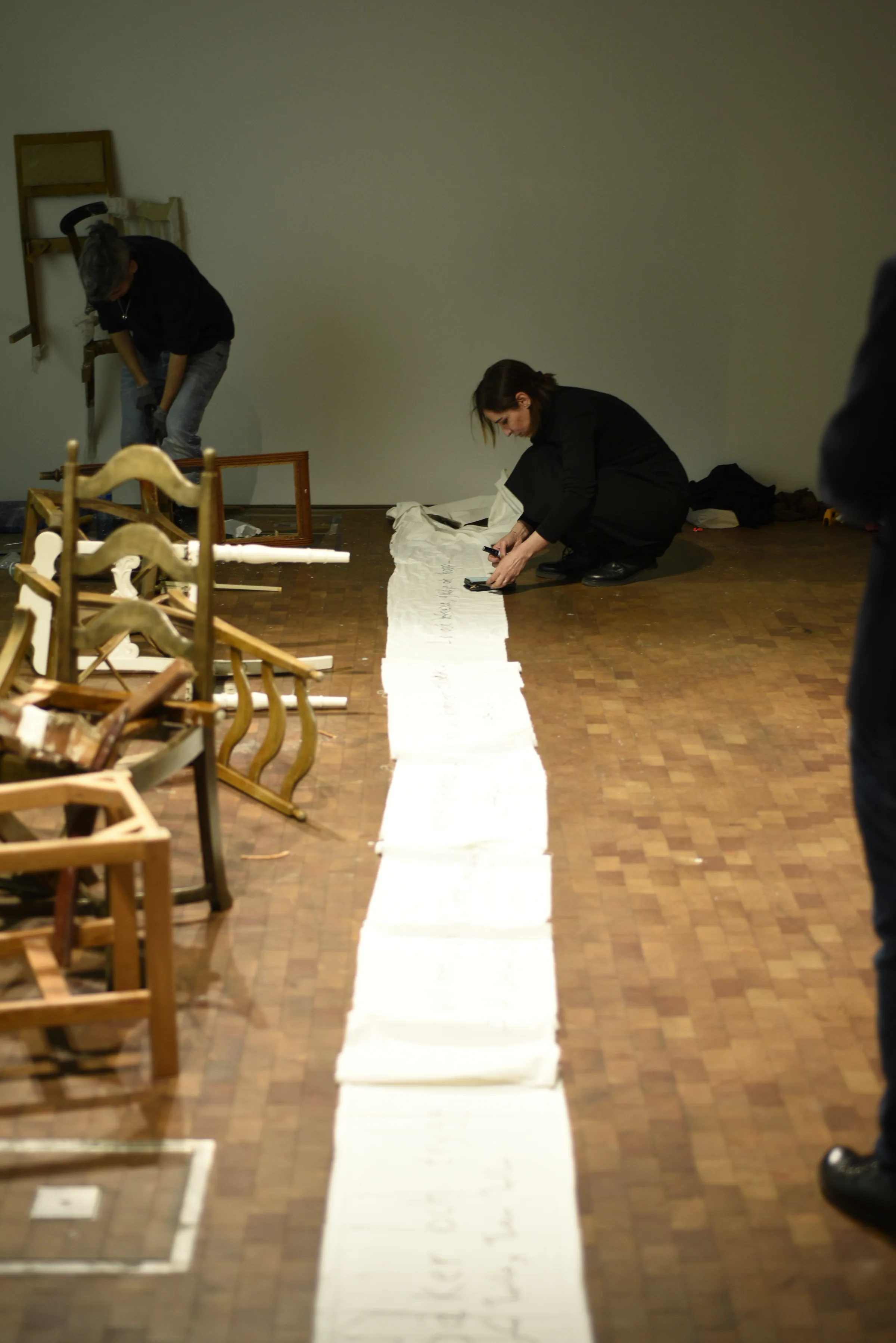 The artist crouches on a wooden floor writing on a long white textile that stretches the length of the room; dismantled wooden chairs are stacked to the left and another person works at the far wall.