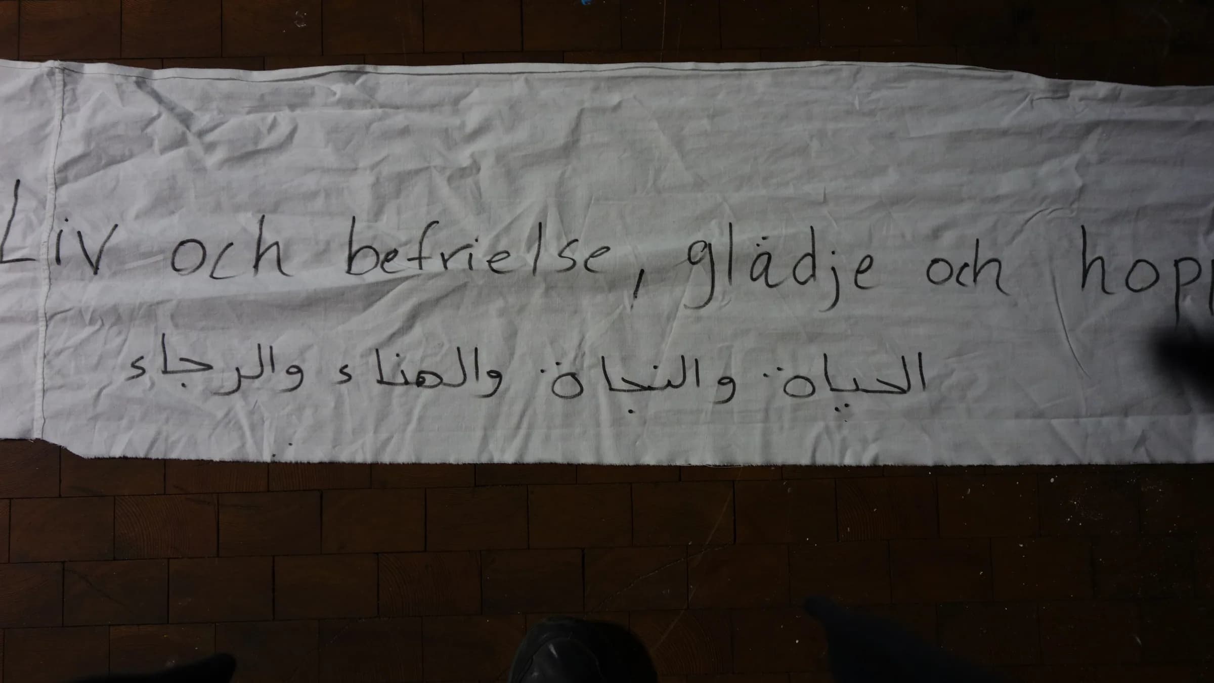 Detail of a long white textile on a wooden floor, handwritten in Swedish “Liv och befrielse, glädje och hopp” with a line of Arabic script beneath it.