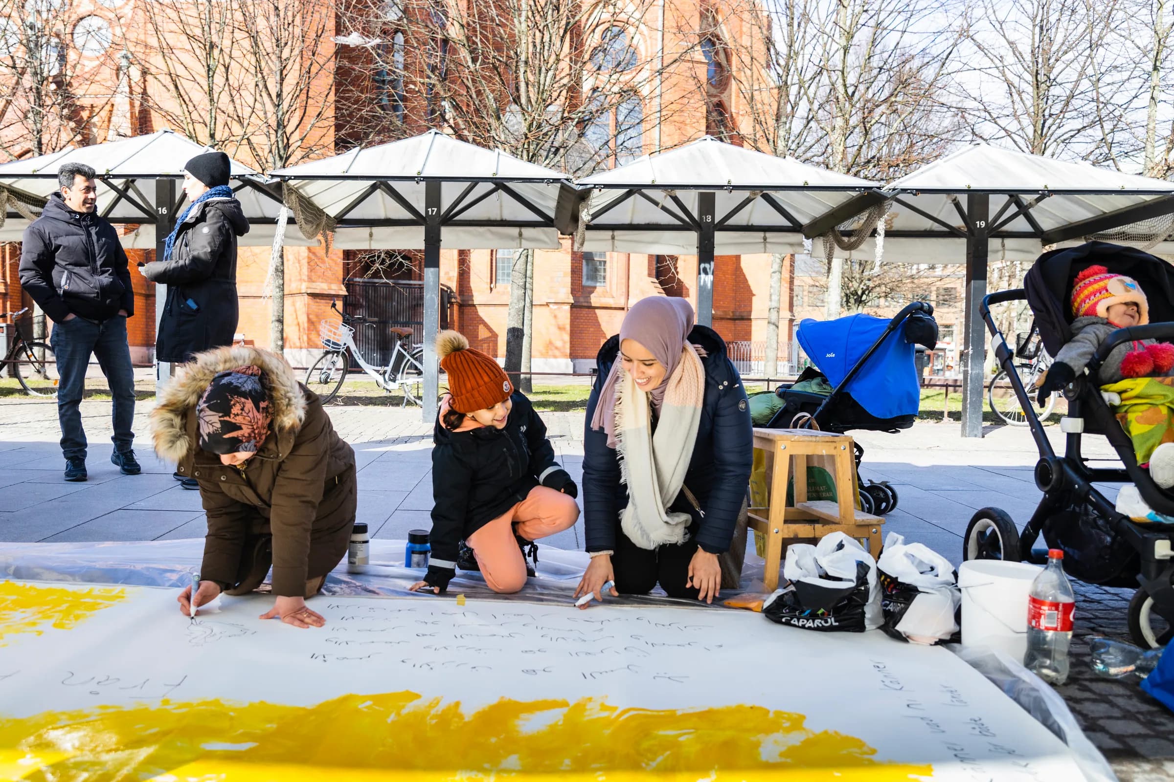 Outdoors on a paved square, a woman in a headscarf and two children kneel over a long white canvas laid on the ground, drawing and writing on it in marker beside a patch of yellow paint. Market stalls and a brick building stand behind them.
