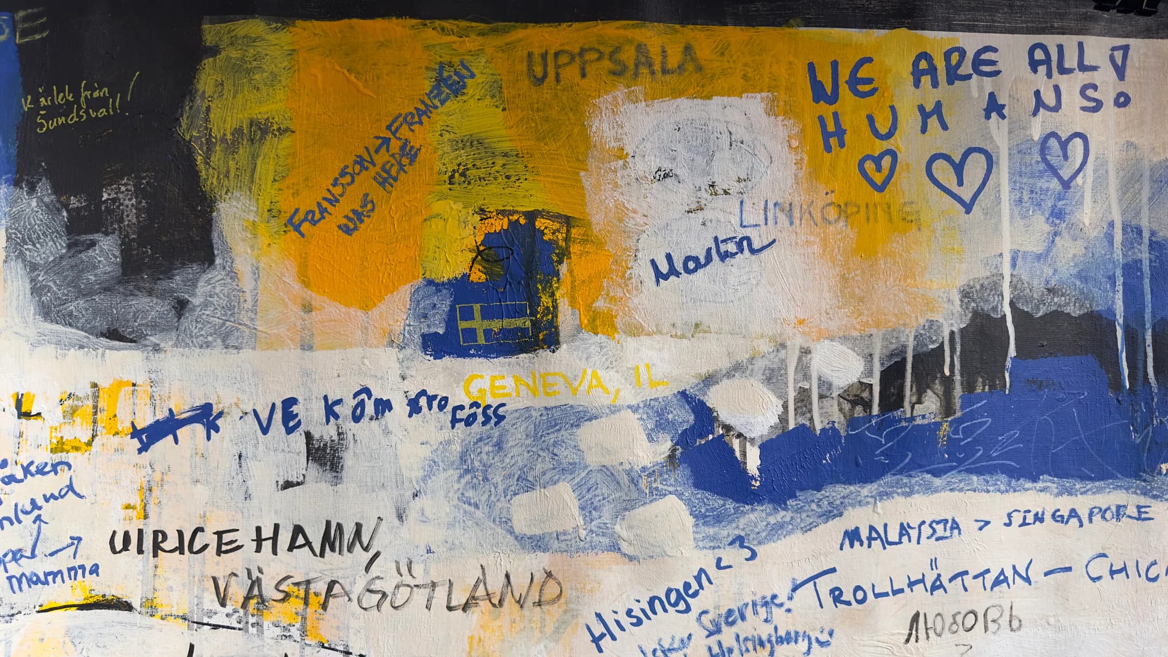 Detail of the finished canvas: layers of yellow, blue and white paint overwritten by hand with place names such as Uppsala and Linköping and messages including “WE ARE ALL HUMANS”.