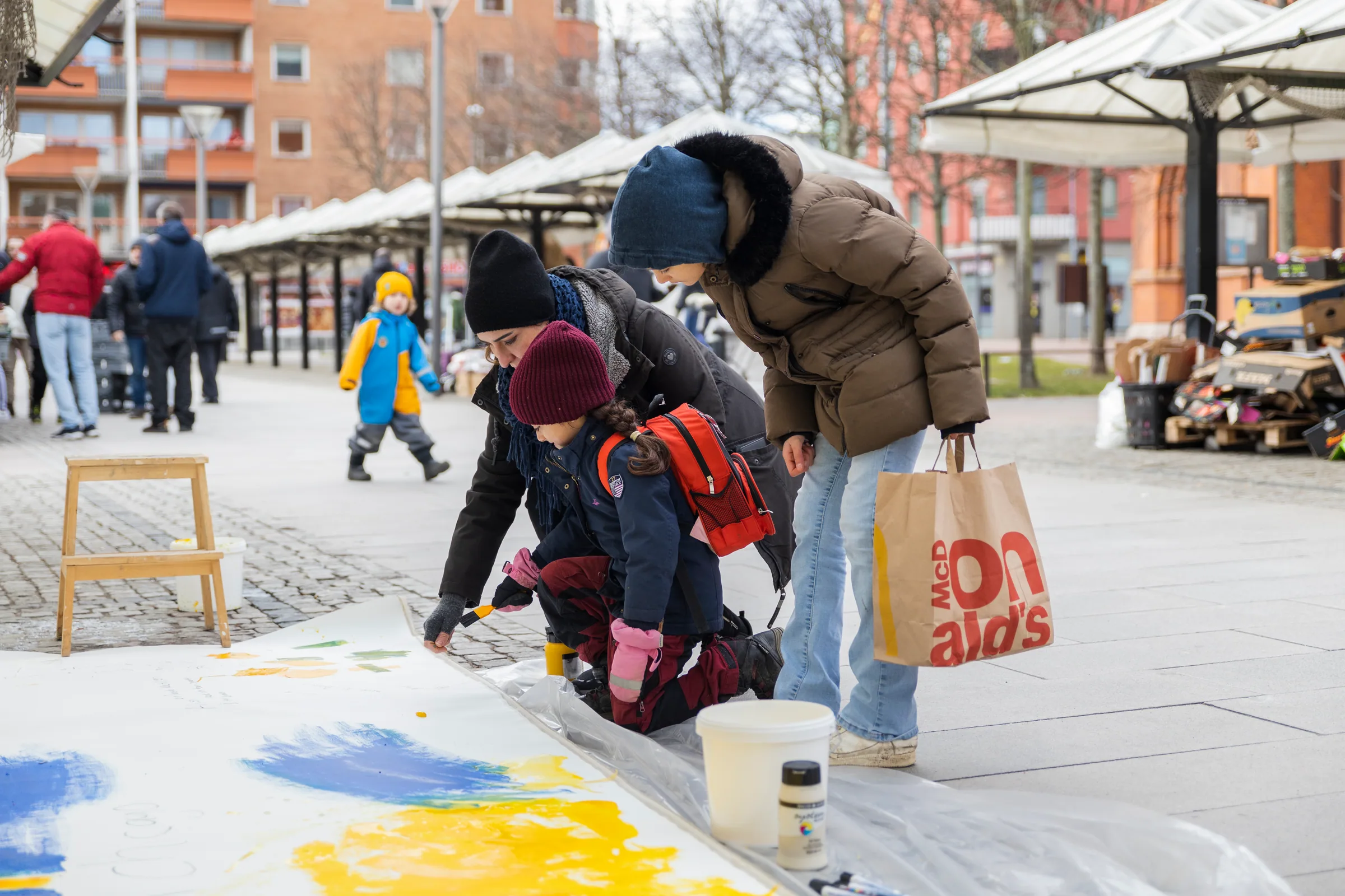 Outdoors, an adult and a small child in winter clothes crouch together to paint yellow and blue marks on a long white canvas on the pavement, while another person watches.