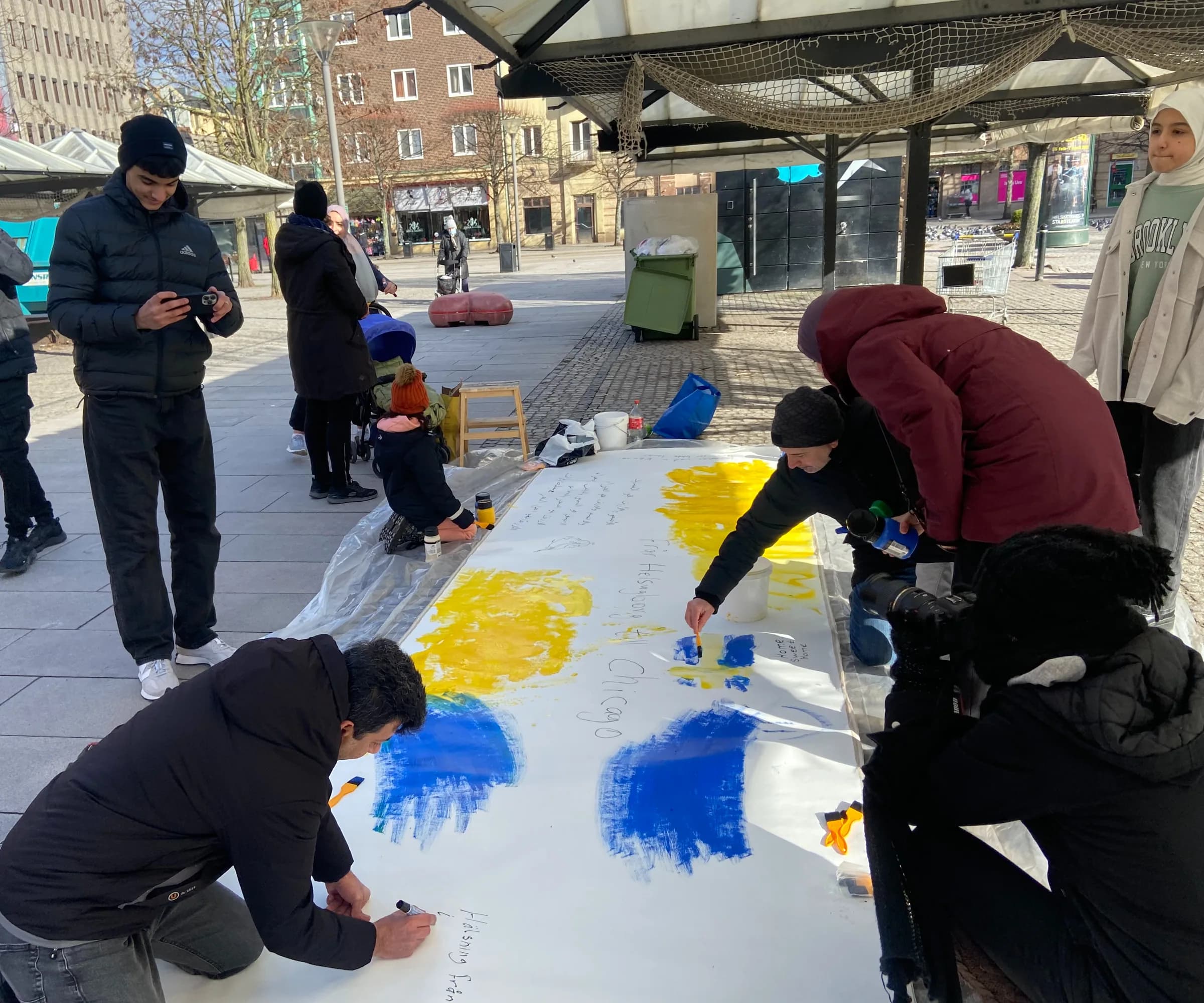 Several people kneel along both sides of a long canvas on a public square, painting broad blue and yellow strokes and writing place names including “Chicago” on the white surface.