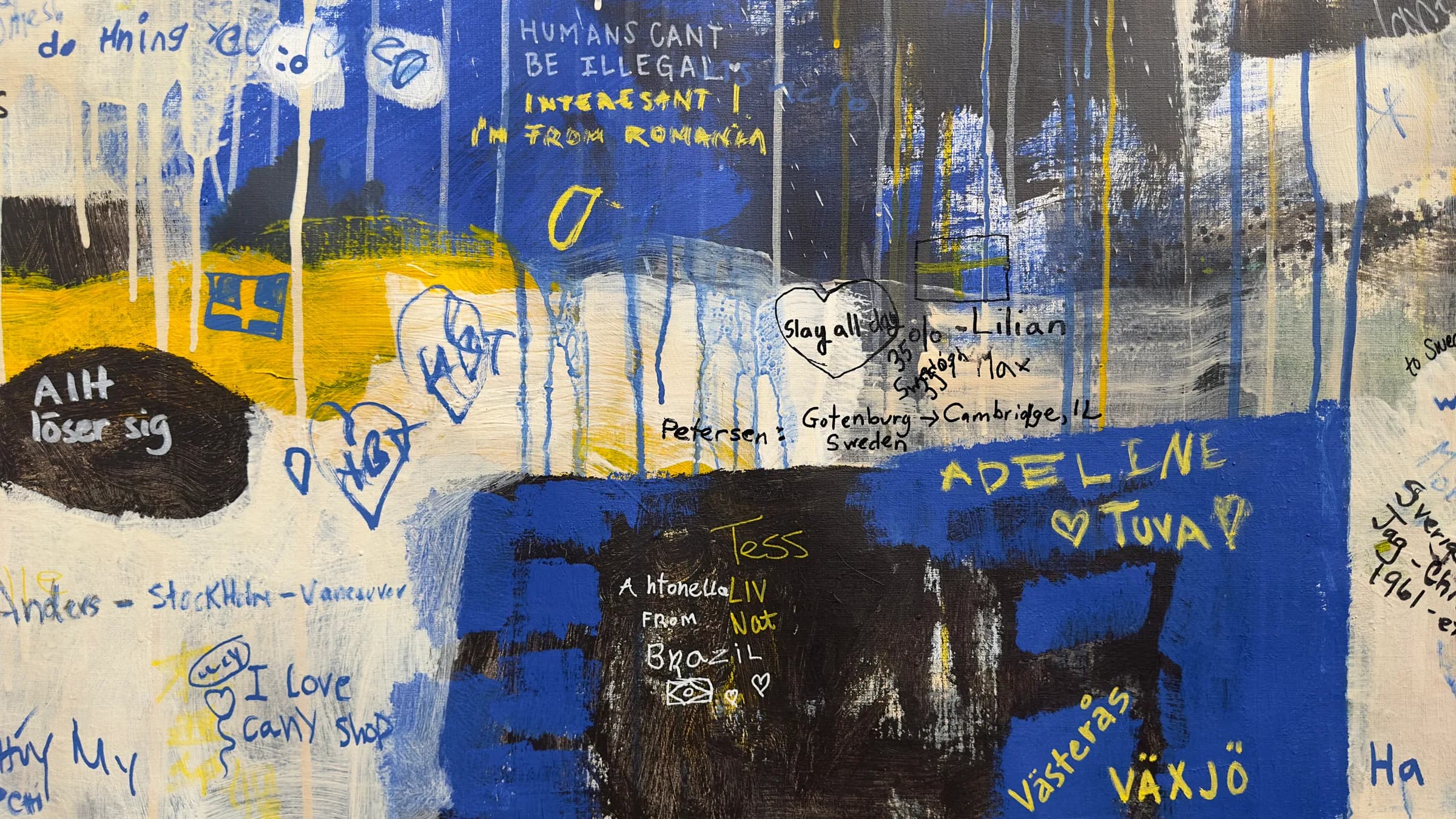 Detail of the finished canvas: blue and yellow paint with a small painted Swedish flag, surrounded by handwritten contributions including “HUMANS CANT BE ILLEGAL” and names of towns and countries.