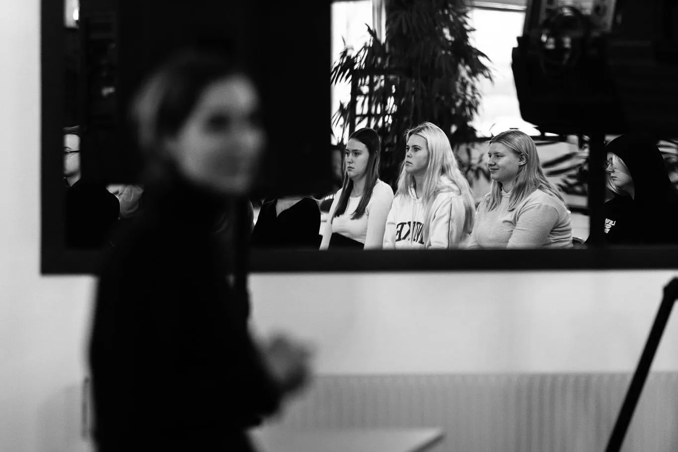 Black-and-white photograph: an out-of-focus figure in black stands in the foreground while a row of seated audience members is reflected in a large mirror behind.