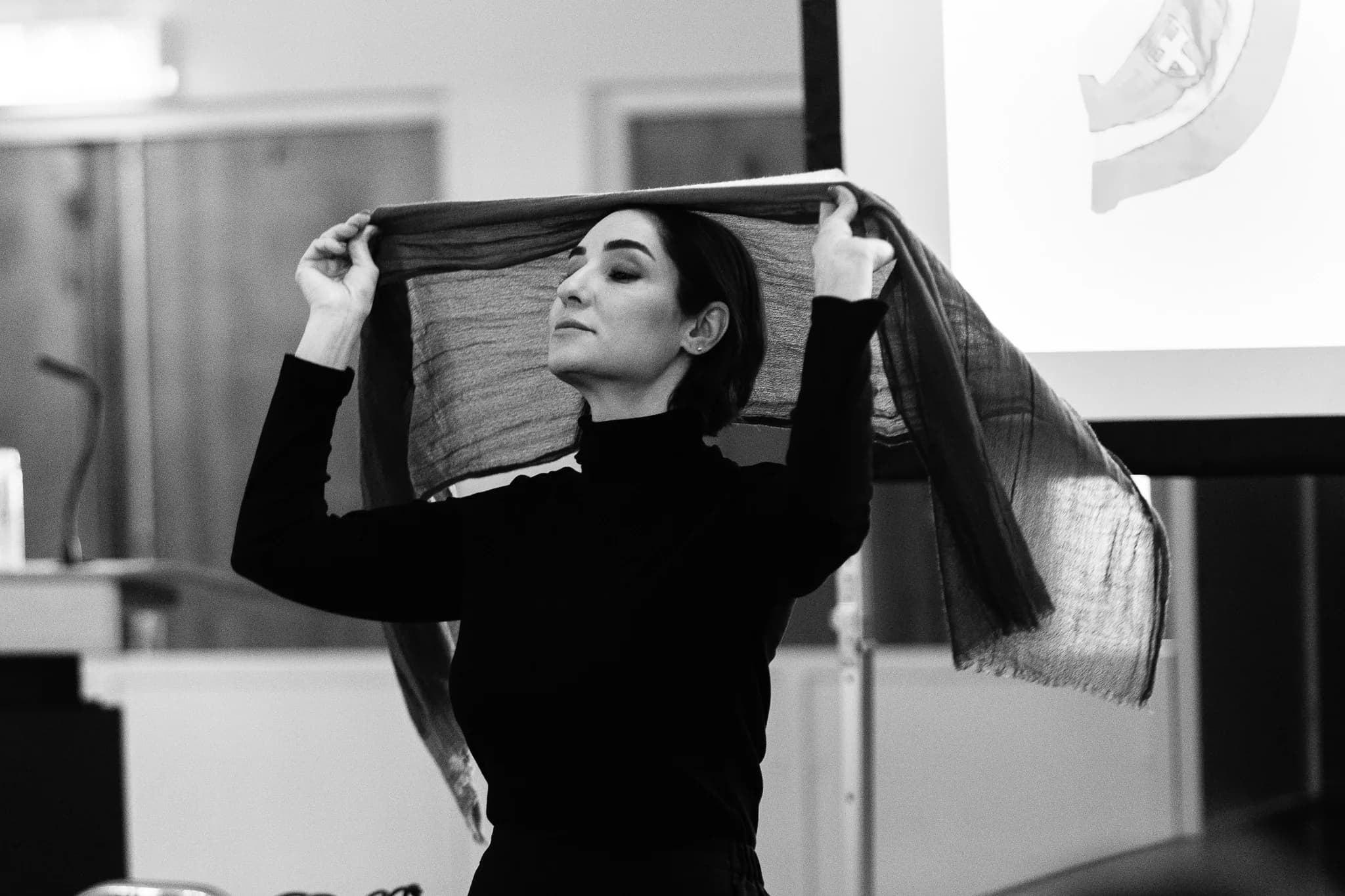 Black-and-white photograph: the artist stands with her eyes closed, lifting a long scarf above and behind her head with both hands, a projection screen visible to her right.