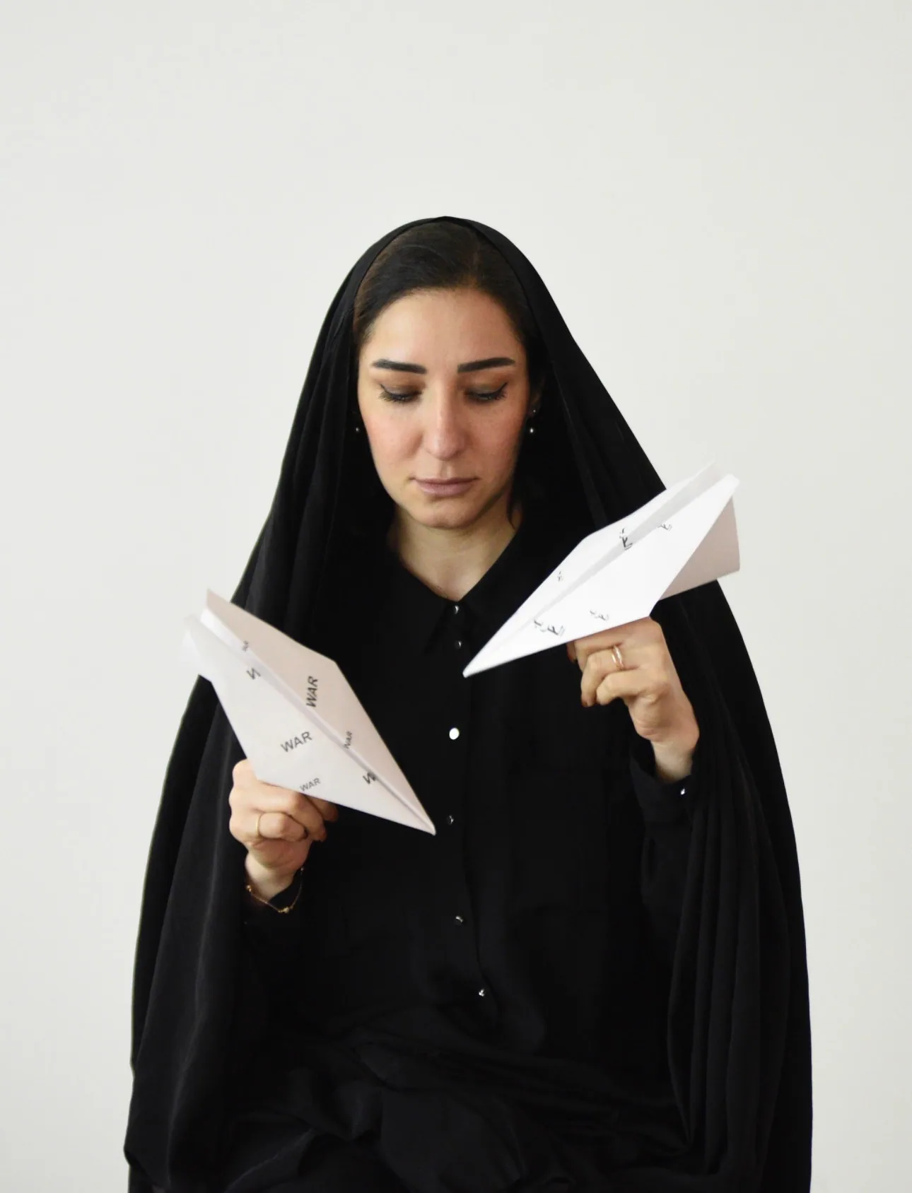 A woman in a black head covering and shirt looks down at two folded paper planes she holds at chest height; the paper is printed with the word “WAR” in Latin and Arabic script, against a plain pale background.