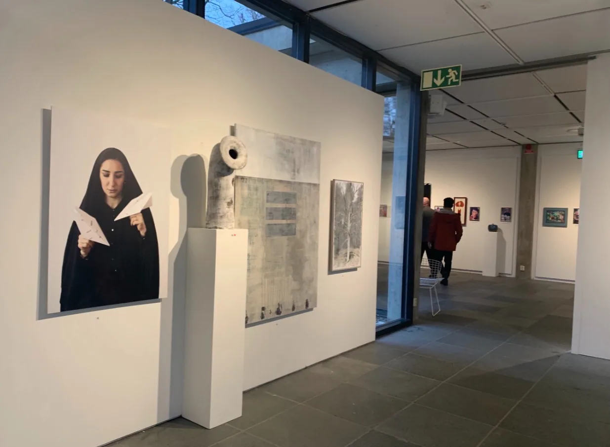 Exhibition view: the Paper Missiles photograph hangs on a white gallery wall beside a sculpture on a plinth and further works, with a visitor walking through the room beyond.
