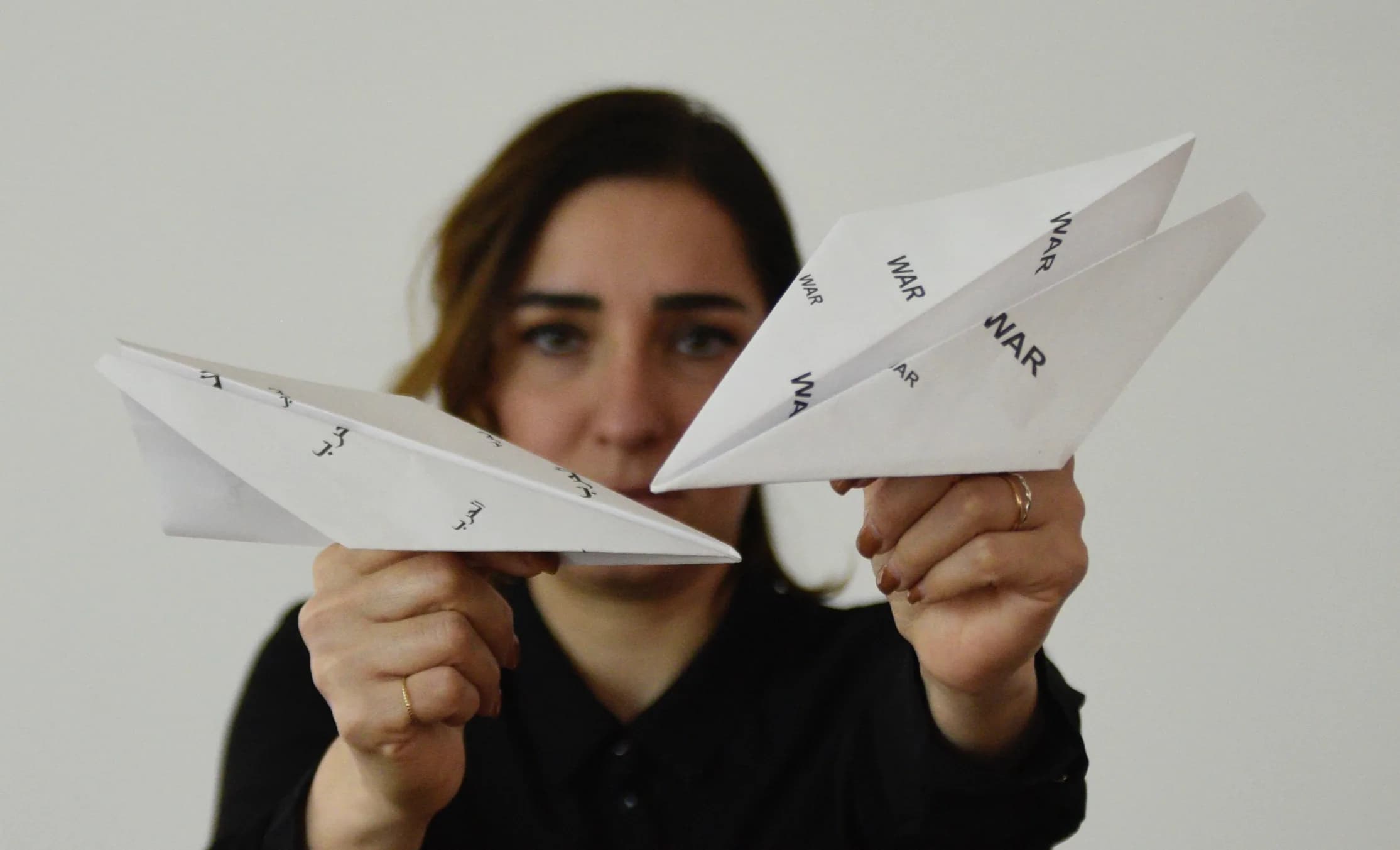 A woman holds up two folded paper planes towards the camera; the paper is printed repeatedly with the word “WAR” in Latin and Arabic script, partly obscuring her face behind them.