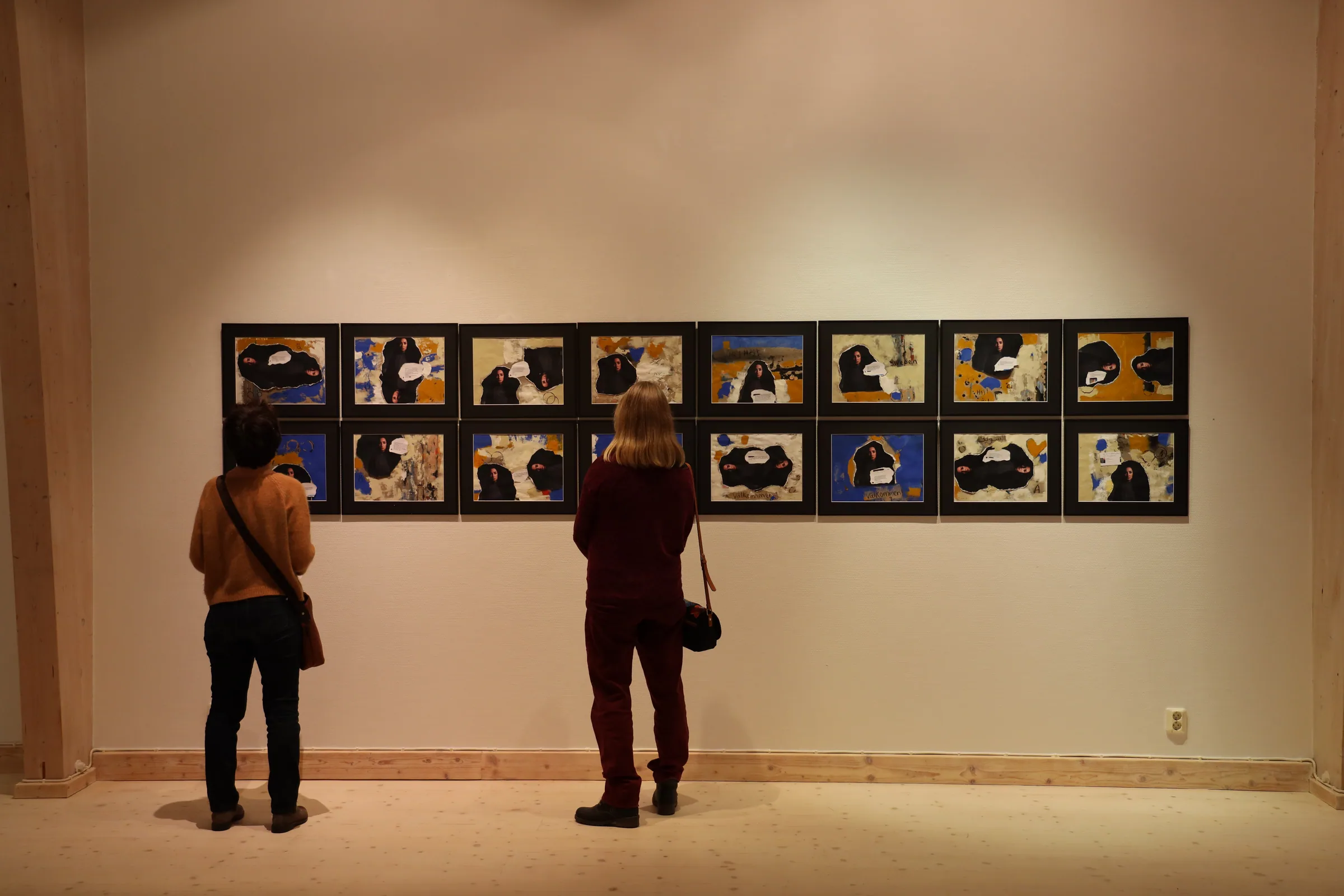 Two visitors stand before a long grid of sixteen framed collages hung in two rows, each combining a cut-out photograph of the artist in a dark veil with blue, yellow and white painted grounds and printed message fragments.