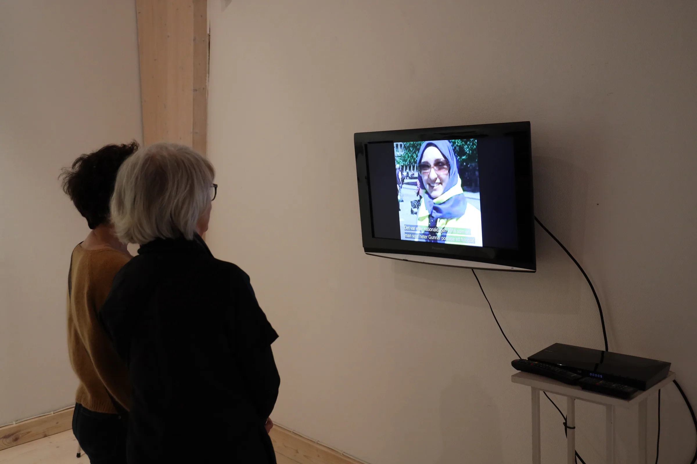 Two visitors watch a wall-mounted screen showing the documentary film, in which a woman in a blue and yellow scarf speaks to camera on a city street, with Swedish subtitles across the lower frame.