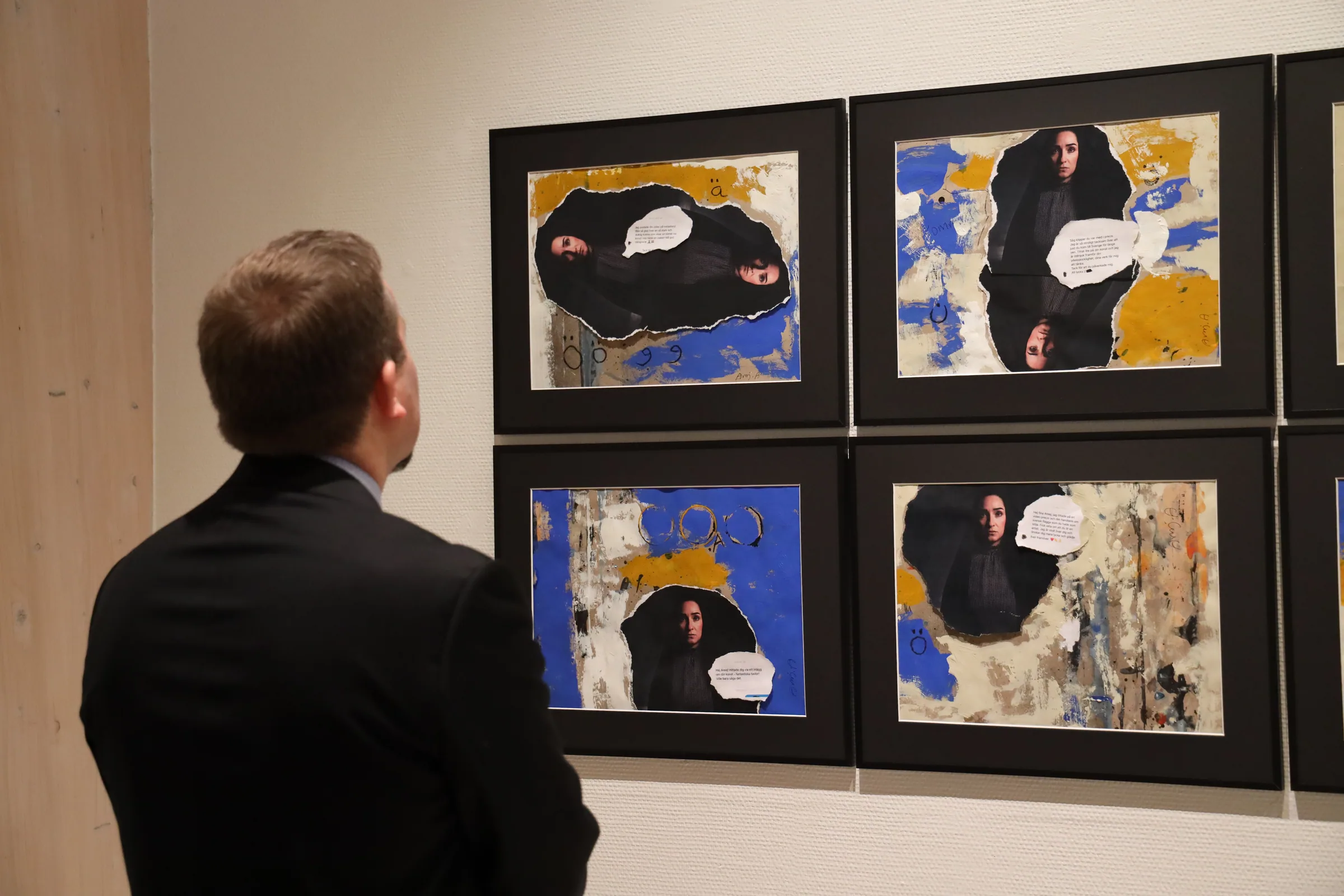 A visitor in a dark suit looks closely at four framed collages, each pairing a torn-out photograph of the artist with painted blue, yellow and white grounds and a printed message in a speech-bubble shape.