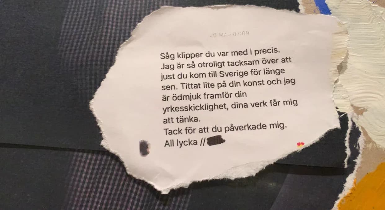 Close-up of a collage panel: a torn paper fragment printed with a longer Swedish message ending “All lycka”, the sender's name blacked out, set against dark checked fabric and pale painted texture.
