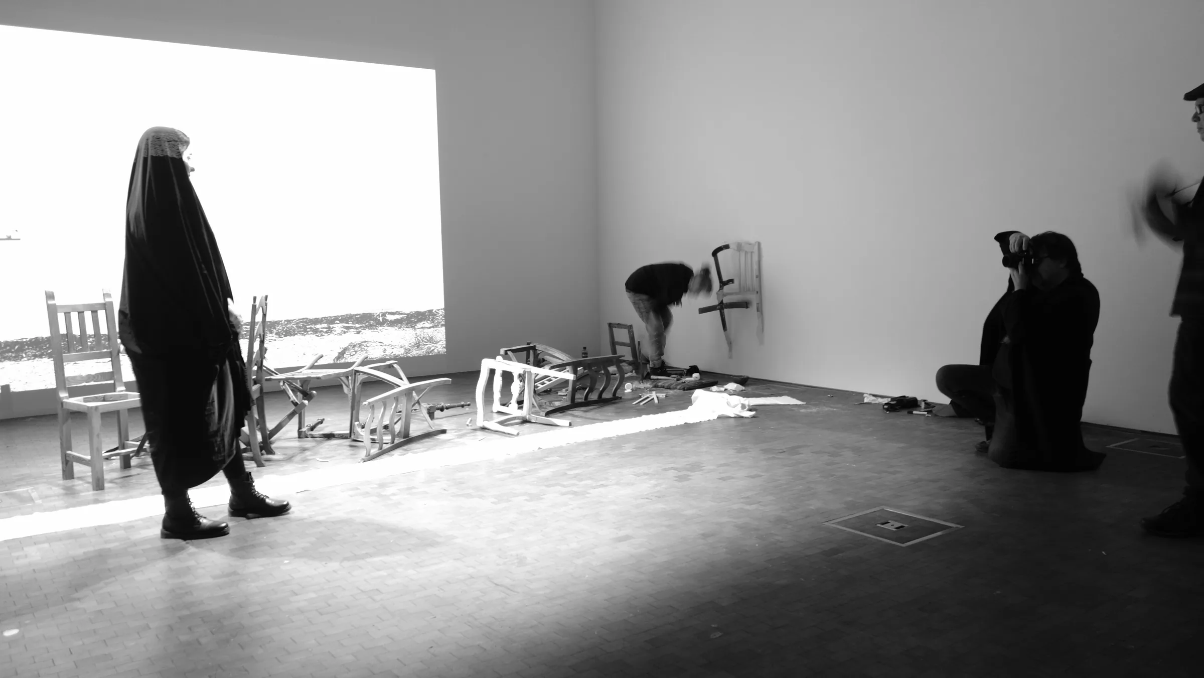 Black-and-white gallery view: a figure draped head to foot in black walks past broken and dismantled chairs scattered on the floor while another person fixes a chair to the wall and a photographer records the scene.