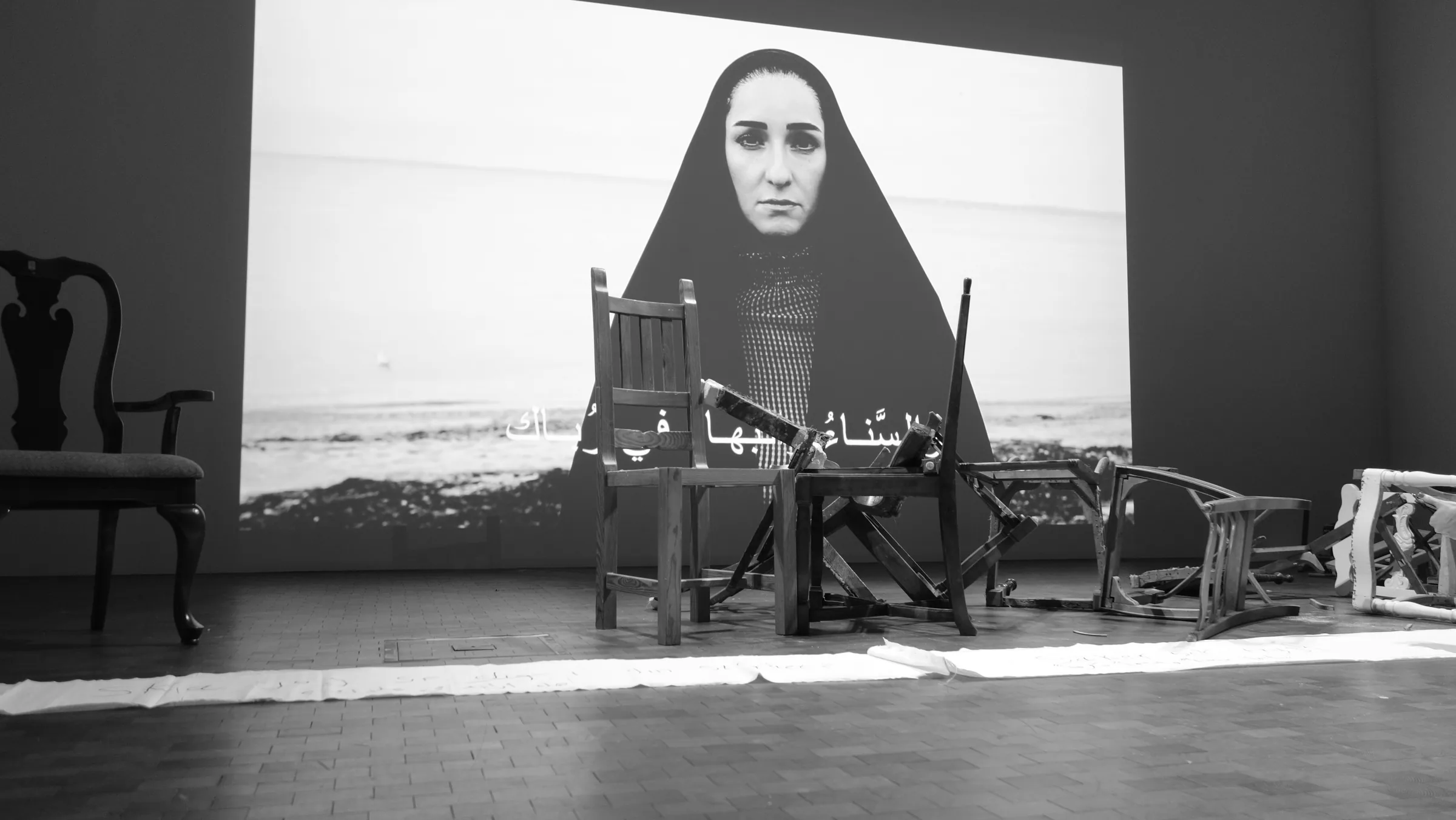 Black-and-white gallery view: a large projection of a woman in a black head covering fills the back wall with Arabic subtitles across it, while wooden chairs stand upright and overturned in front of the screen.