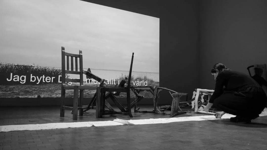 Black-and-white view of a gallery where dismantled wooden chairs stand and lie in front of a wall projection; a projected Swedish line reads “Jag byter Dig … mot allt i … värld” and a figure crouches at the right.