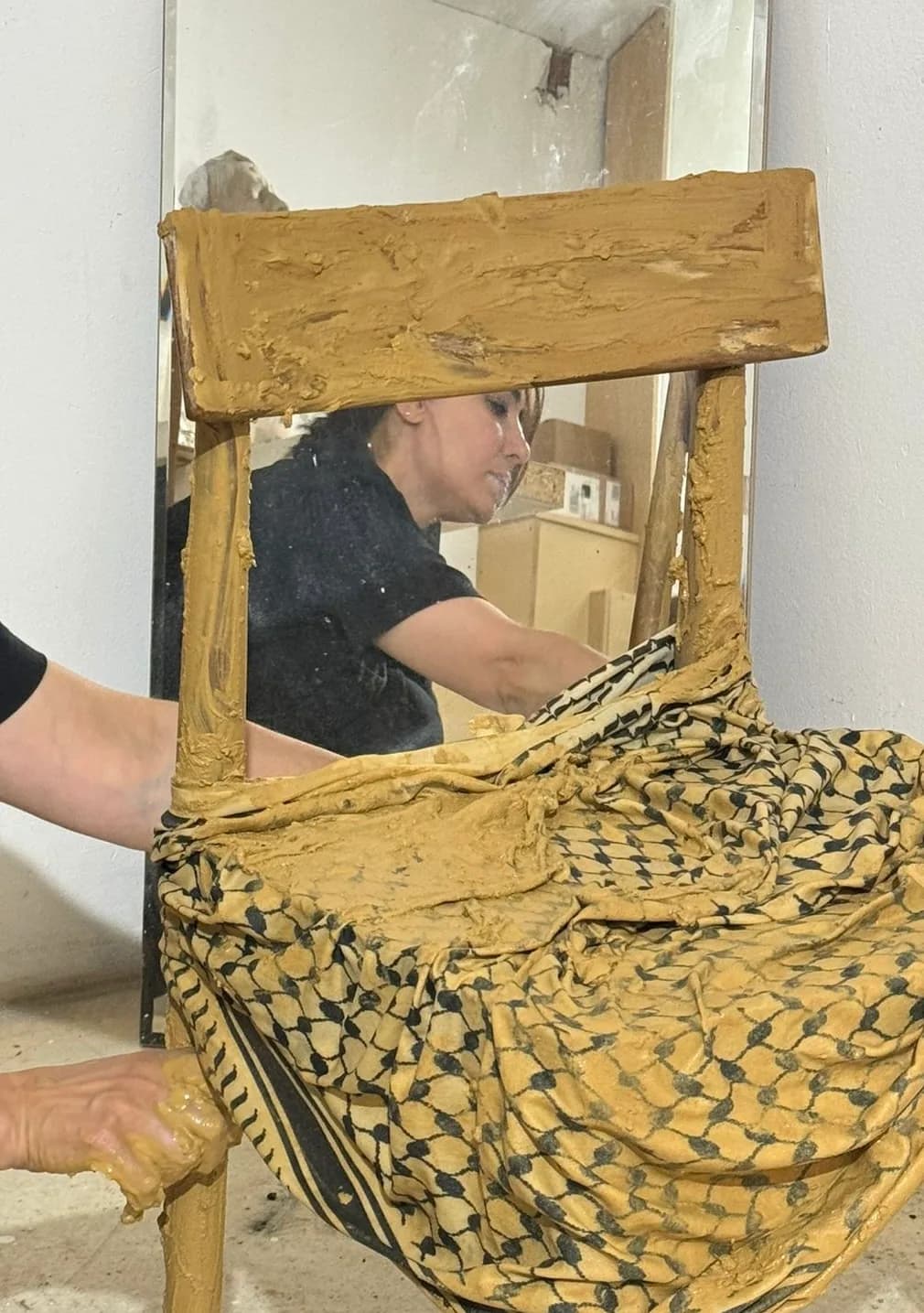 A wooden chair draped with a patterned cloth is being coated in ochre clay by hand; the artist is visible working in a mirror leaning against the wall behind it.