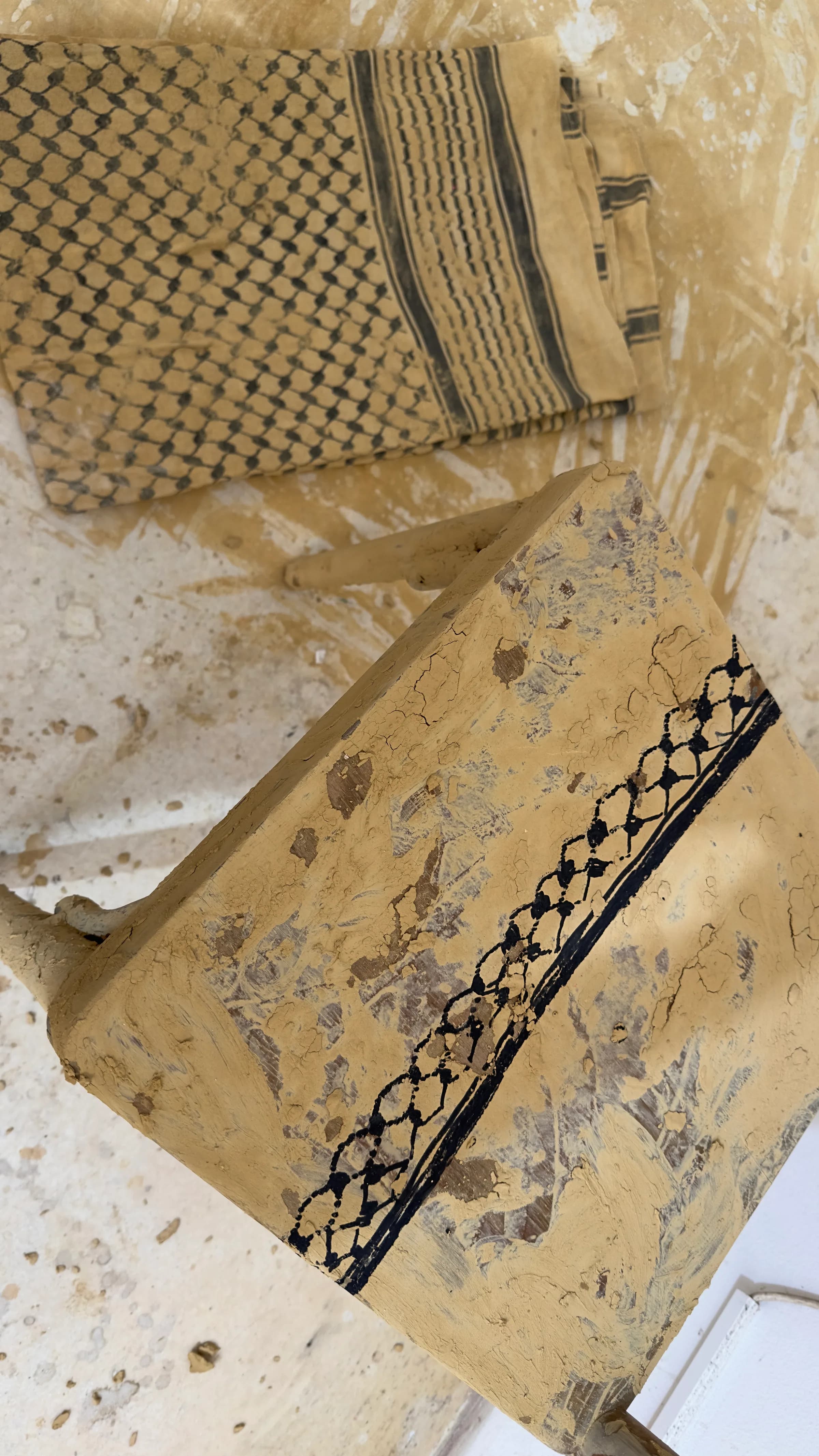 Seen from above, a folded patterned cloth stiffened with dried ochre clay lies beside a clay-covered chair seat on a clay-spattered floor.