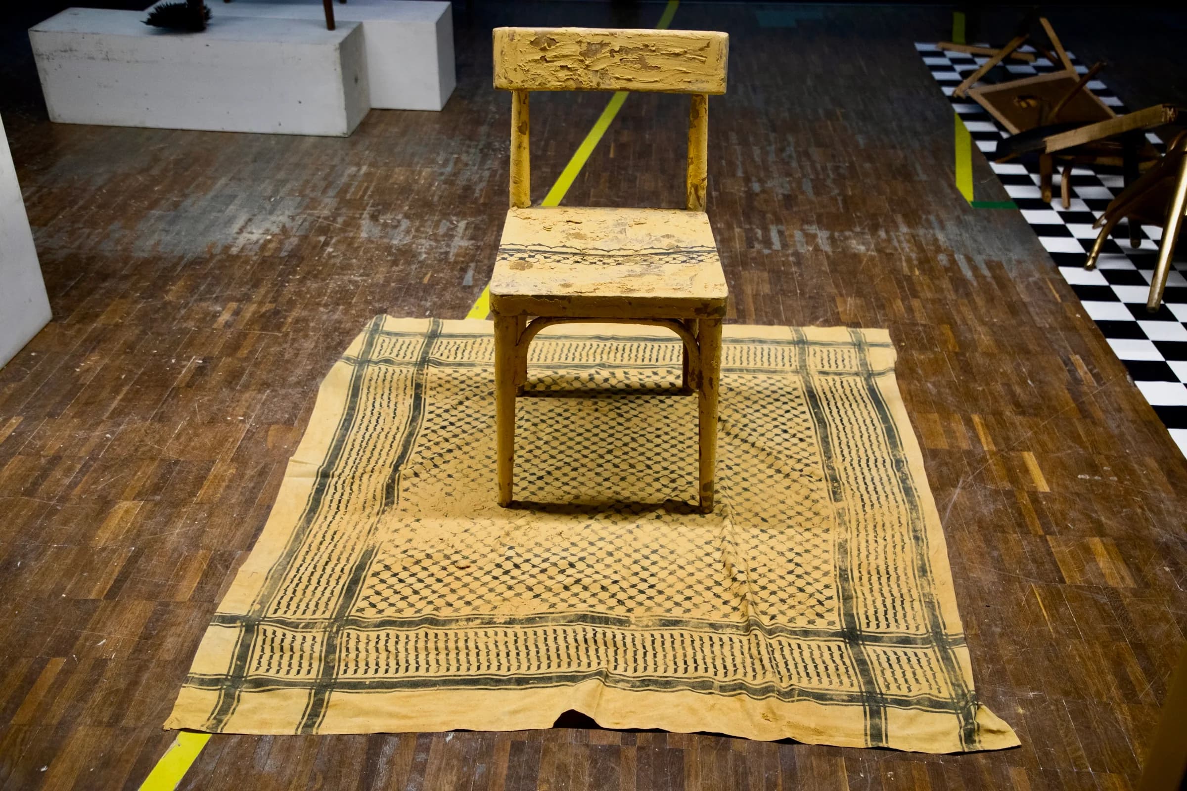 Installation view: a chair entirely coated in dried ochre clay stands on a clay-soaked patterned cloth laid across a wooden floor, with overturned chairs on a checkerboard surface to the right.