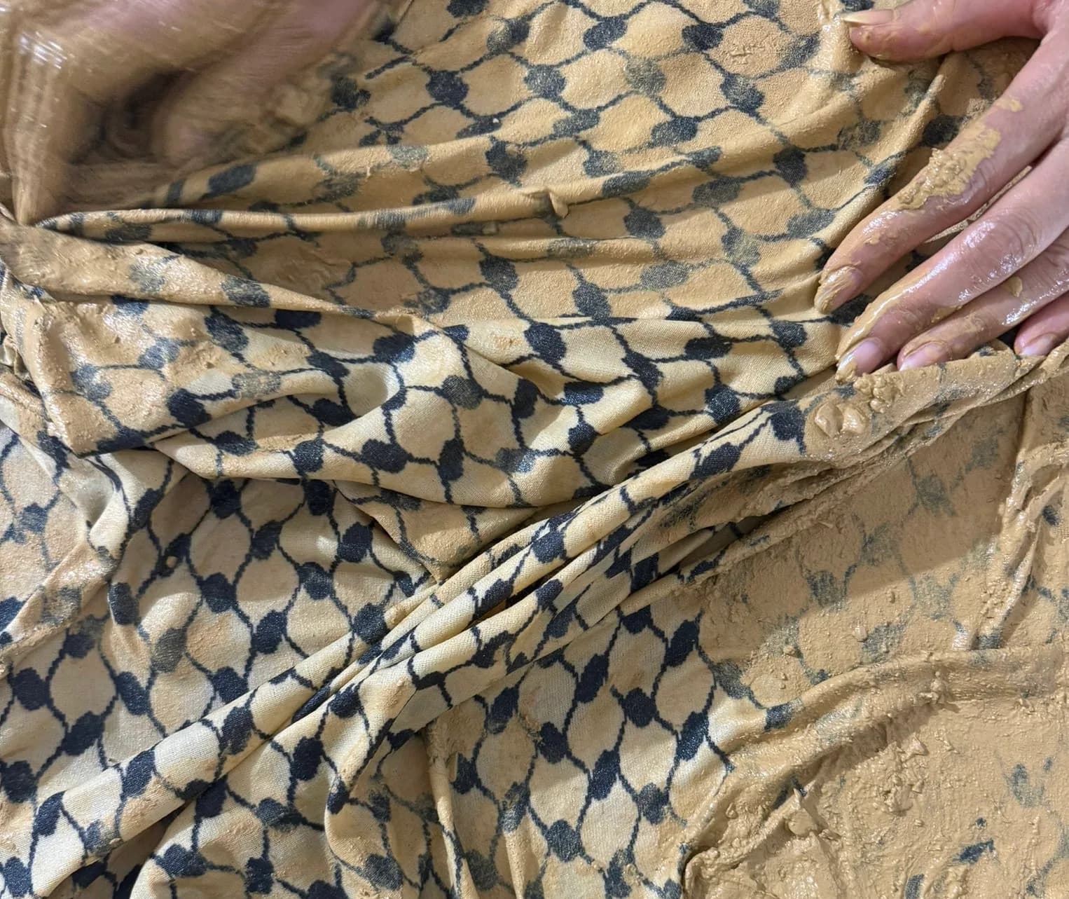 Close-up of two hands pressing wet ochre clay into a black-and-white patterned cloth, the fabric partly absorbed into the clay surface.