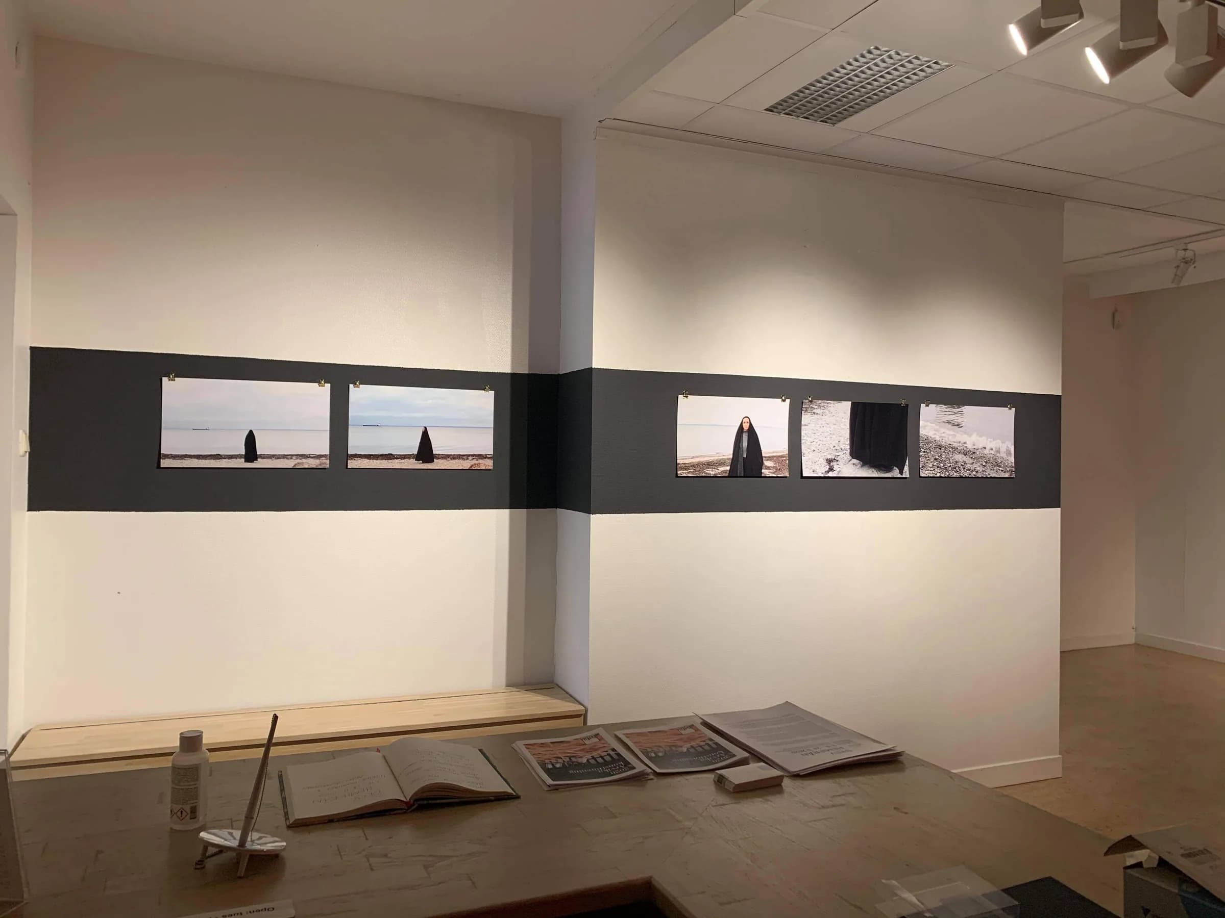 Installation view: five panoramic photographs of a cloaked figure by the sea are clipped in a row along a dark painted band running across two gallery walls, with an open guest book and newspapers on a table in the foreground.