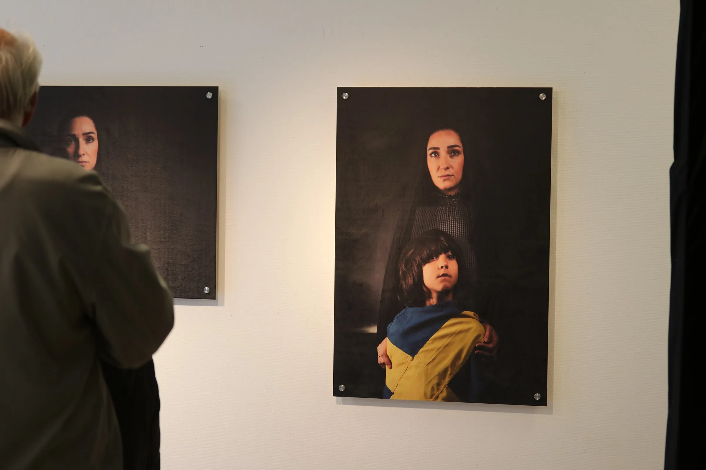 Two mounted photographic works on a gallery wall: one shows a woman in a dark veil, the other shows the same woman standing behind a child wrapped in a blue and yellow Swedish flag, her hands on the child's shoulders. A visitor looks on from the left.