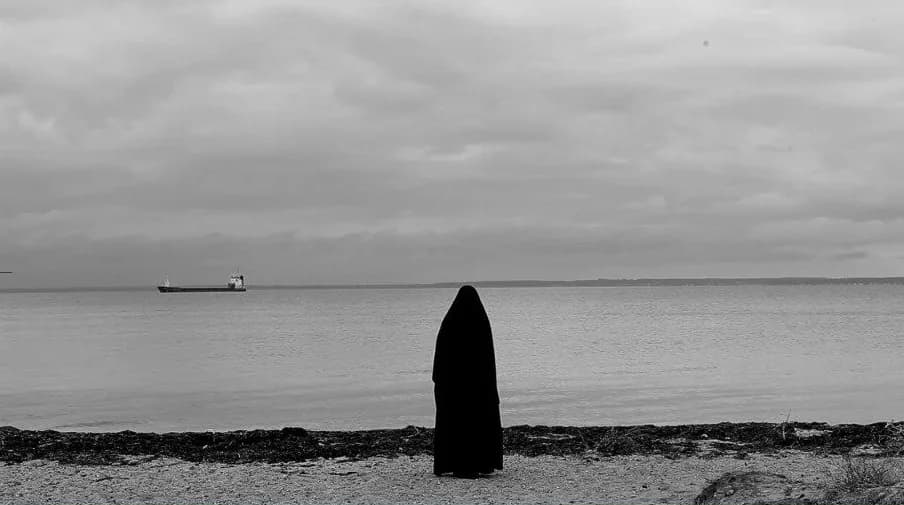 Black-and-white photograph: a lone figure wrapped in a long black cloak stands on a pebble shore facing a calm sea, with a cargo ship on the horizon under an overcast sky.