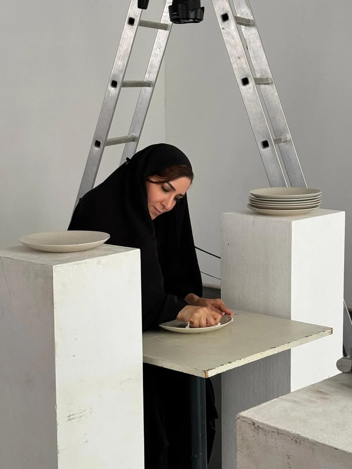 A woman in a black abaya and headscarf leans over a plate resting on a white plinth; a stack of plain plates sits on a second plinth and a stepladder with a camera mounted on it stands behind her.