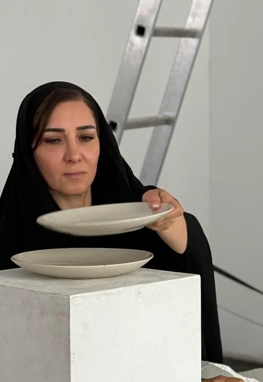 The artist in black holds one plain plate raised above another that rests on a white plinth, looking down at them.