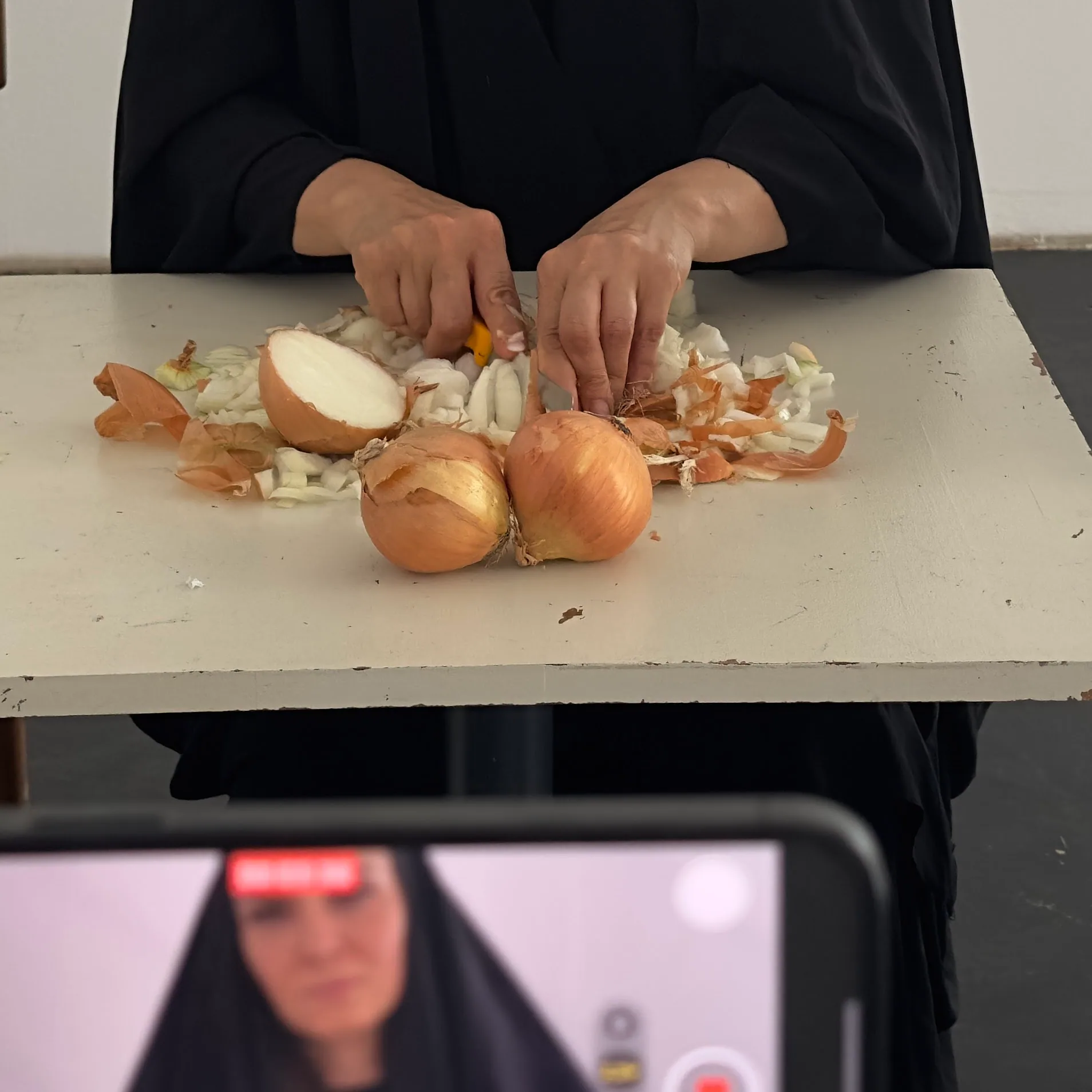 Two hands cut onions on a white table strewn with skins and chopped pieces; a phone on a stand in the foreground records the action.
