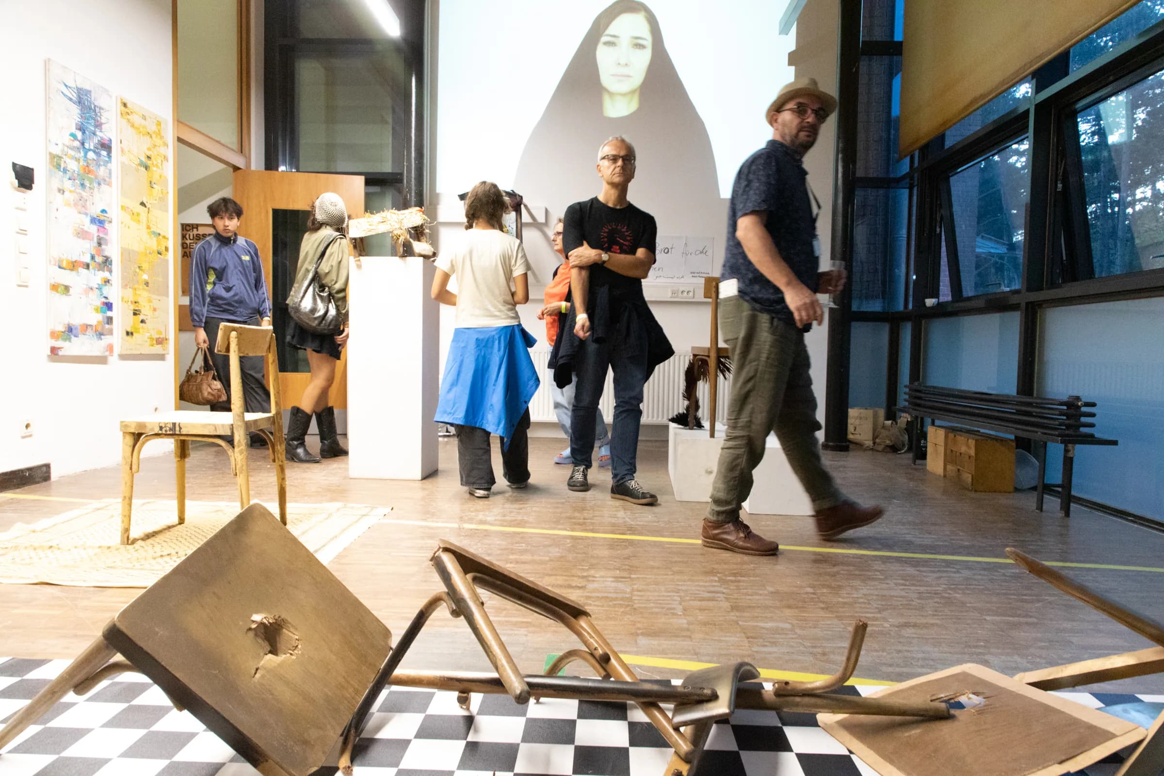 Exhibition view: visitors move through a room where wooden chairs lie broken across a black-and-white checkerboard floor, in front of a large projection of a woman in a black head covering.